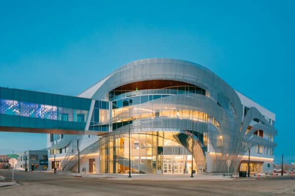 Curved glass-and-metal civic building glows internally, its sweeping exterior ribbons framing a multi-story atrium; a glass skybridge connects to another block at a quiet urban intersection at dusk.

Visible text: "Bettie Cram" (street sign); "SEE YOU" (on building).