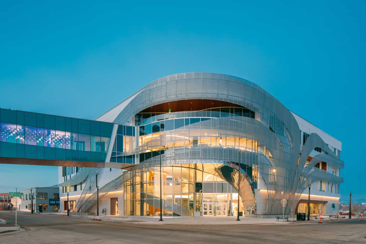 Curved glass-and-metal civic building glows internally, its sweeping exterior ribbons framing a multi-story atrium; a glass skybridge connects to another block at a quiet urban intersection at dusk.

Visible text: "Bettie Cram" (street sign); "SEE YOU" (on building).