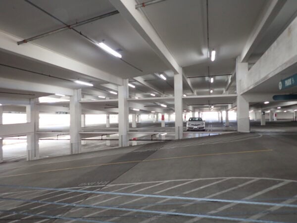 A white sedan sits parked between concrete columns in a mostly empty multi-level parking garage, fluorescent lights overhead and striped ramps and lanes running through the spacious interior.
