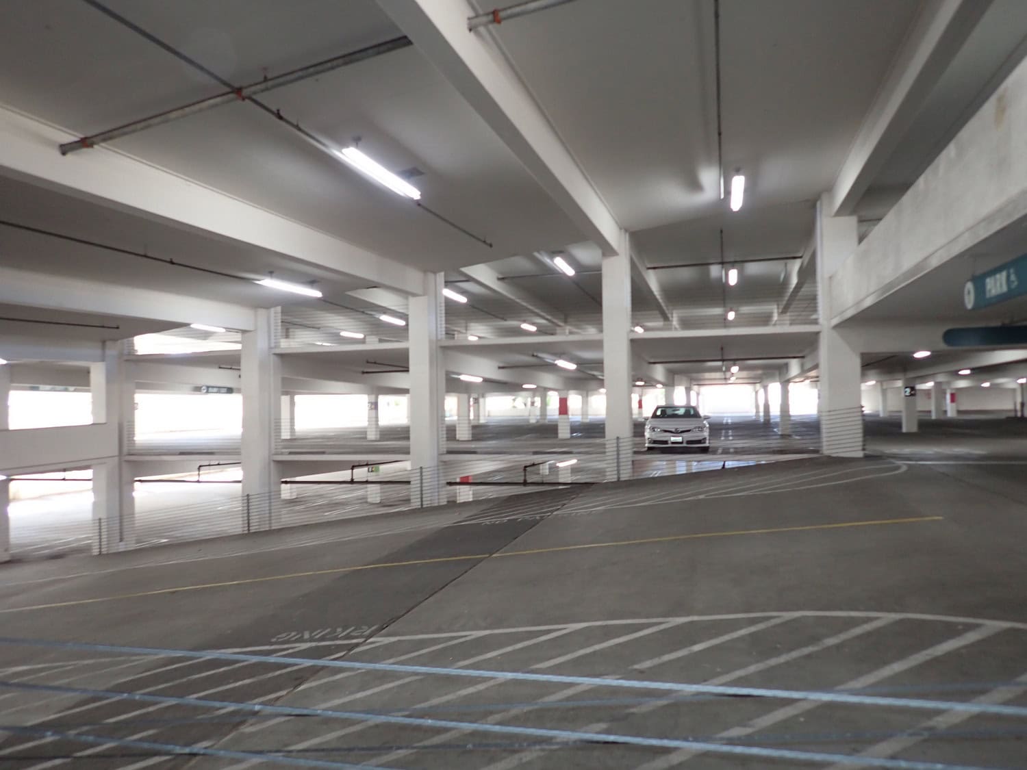 A white sedan sits parked between concrete columns in a mostly empty multi-level parking garage, fluorescent lights overhead and striped ramps and lanes running through the spacious interior.