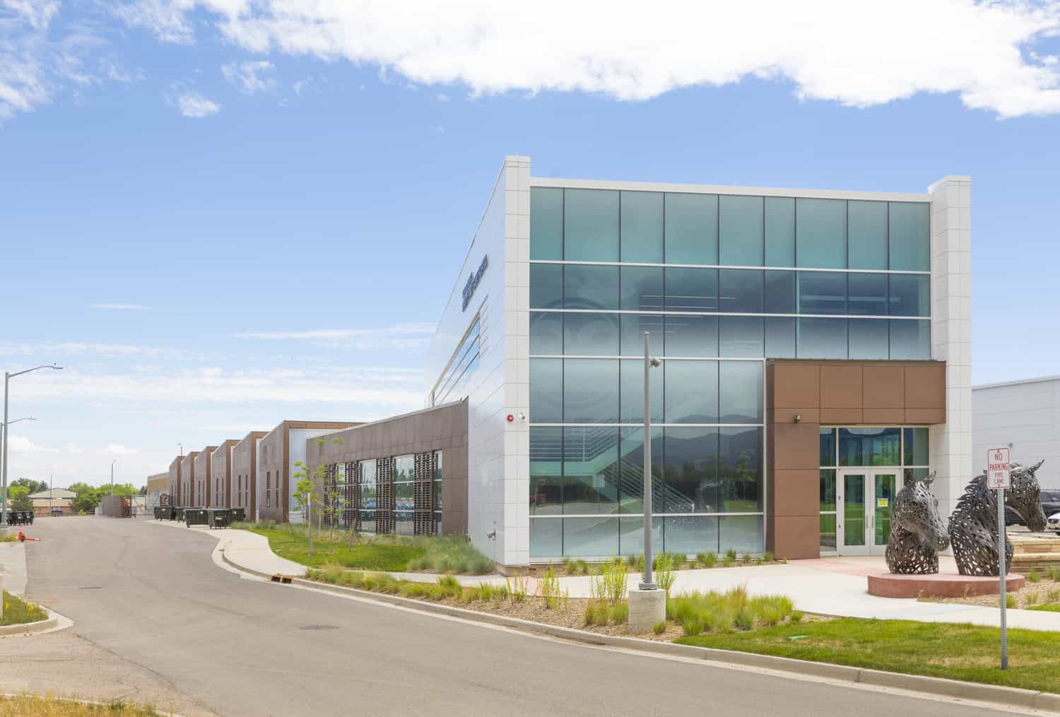 Glass-and-metal building fronts a quiet road; two woven-metal horse sculptures sit by the entrance, minimal landscaping and a curved street beside it under blue sky. Text: "NO PARKING FIRE LANE".