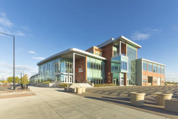 Modern multi-story glass-and-brick building labeled "CSU SPUR VIDA" stands bathed in sunlight, its floor-to-ceiling windows reflecting a clear blue sky, beside a wide concrete plaza and empty street.