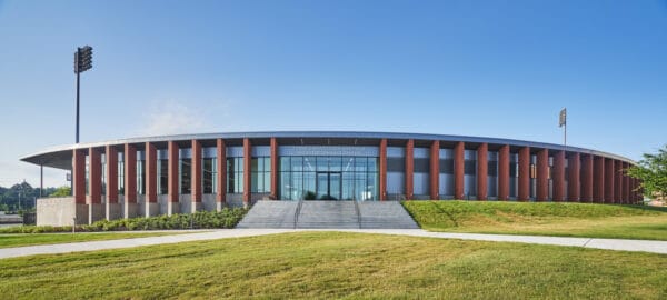 FRANK O'MARA TRACK AND FIELD HIGH PERFORMANCE CENTER — oval brick-and-glass building with vertical columns, glass entrance and broad stairs, set on a manicured lawn with stadium lights under clear blue sky.