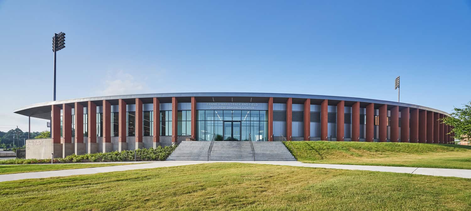 FRANK O'MARA TRACK AND FIELD HIGH PERFORMANCE CENTER — oval brick-and-glass building with vertical columns, glass entrance and broad stairs, set on a manicured lawn with stadium lights under clear blue sky.