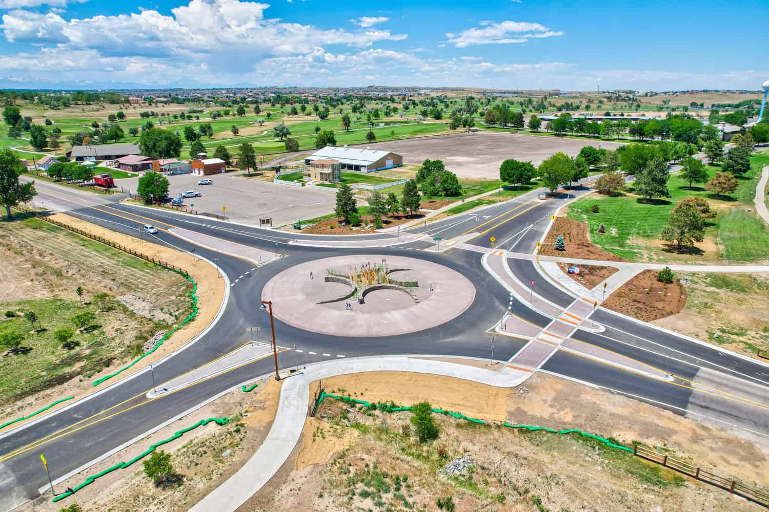 A landscaped roundabout with central sculptural planters channels traffic through four asphalt roads, surrounded by parking lots, scattered buildings, golf-course greens and open fields under a blue, partly cloudy sky.