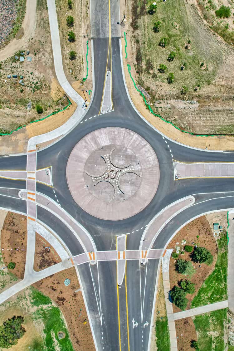 Central circular roundabout guiding vehicular traffic, with painted lanes and splitter islands directing flows; four approach roads connect in a grassy, partially landscaped suburban area with sidewalks.