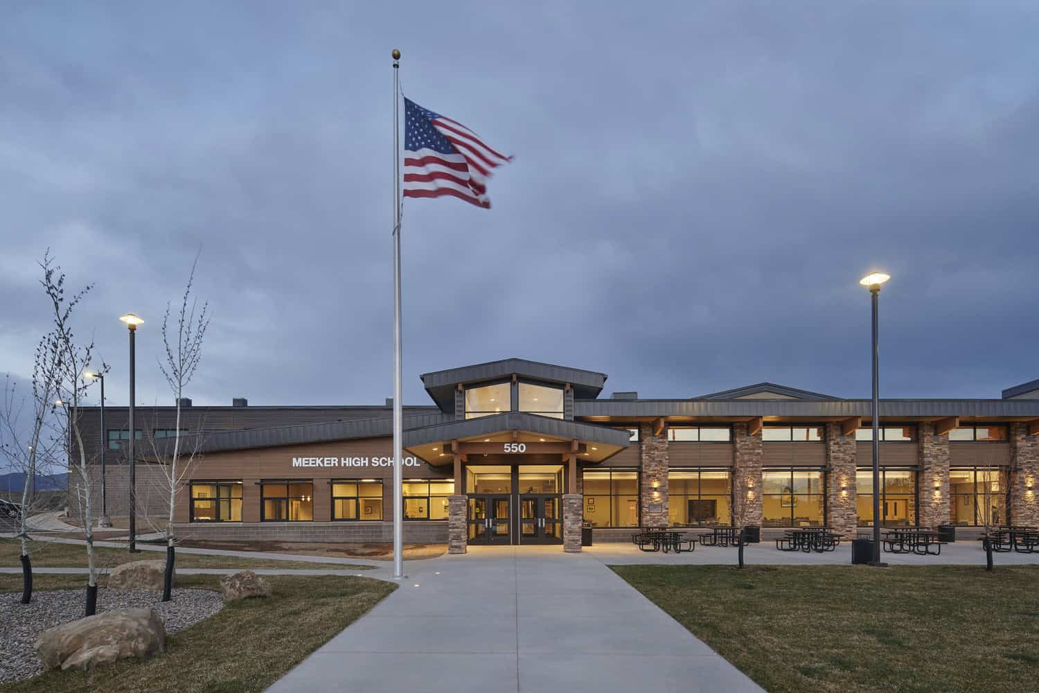 American flag waves on a tall pole above a school entrance; modern two-story building with lit windows, concrete walkway and lampposts at dusk. Text: "MEEKER HIGH SCHOOL", "550".