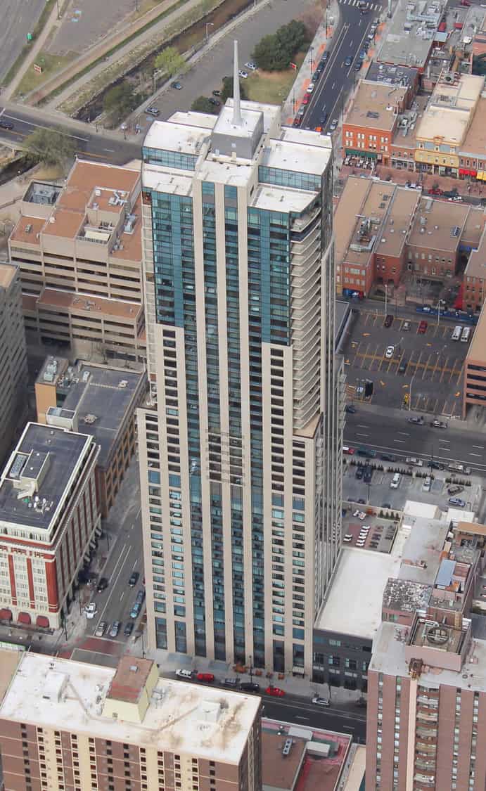 A glass-and-stone high-rise tower rises above downtown, its vertical blue-tinted windows and rooftop spire flanked by balconies, surrounded by shorter commercial buildings, streets, parked cars and surface lots.