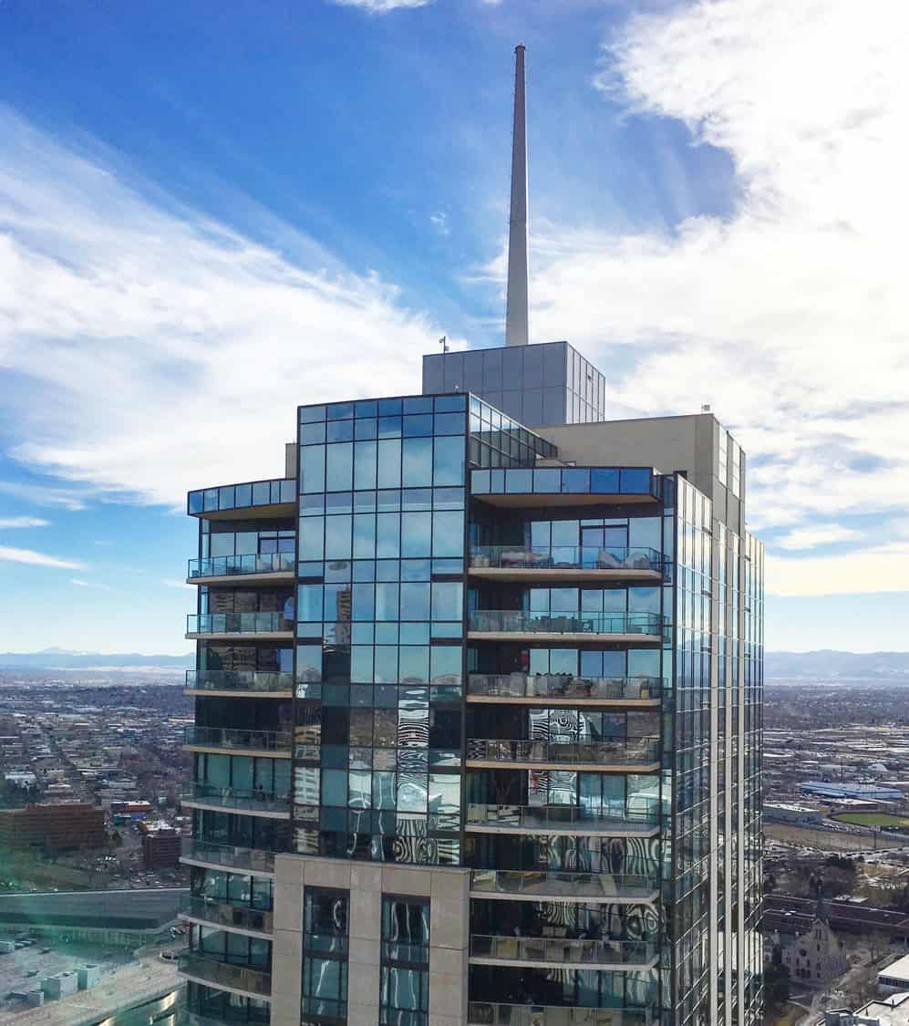 Glass-clad high-rise topped by a tall spire reflects blue sky and surrounding city, with balconies visible, set against a sprawling urban landscape and distant mountains.