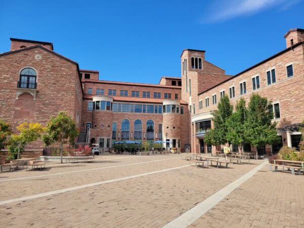 Large red‑brick academic building faces a paved courtyard; arched windows and towers frame tables, benches, and trees, while a person walks near benches under a clear blue sky.