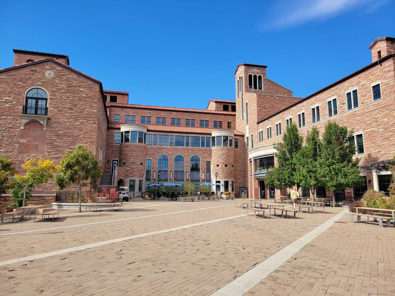 Large red‑brick academic building faces a paved courtyard; arched windows and towers frame tables, benches, and trees, while a person walks near benches under a clear blue sky.