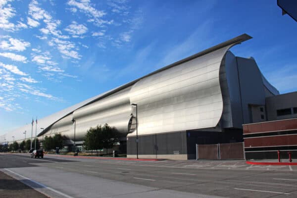 Large curved metallic building stretches along a quiet city street, its smooth silver panels catching sunlight under a bright blue sky with scattered clouds and a few trees.