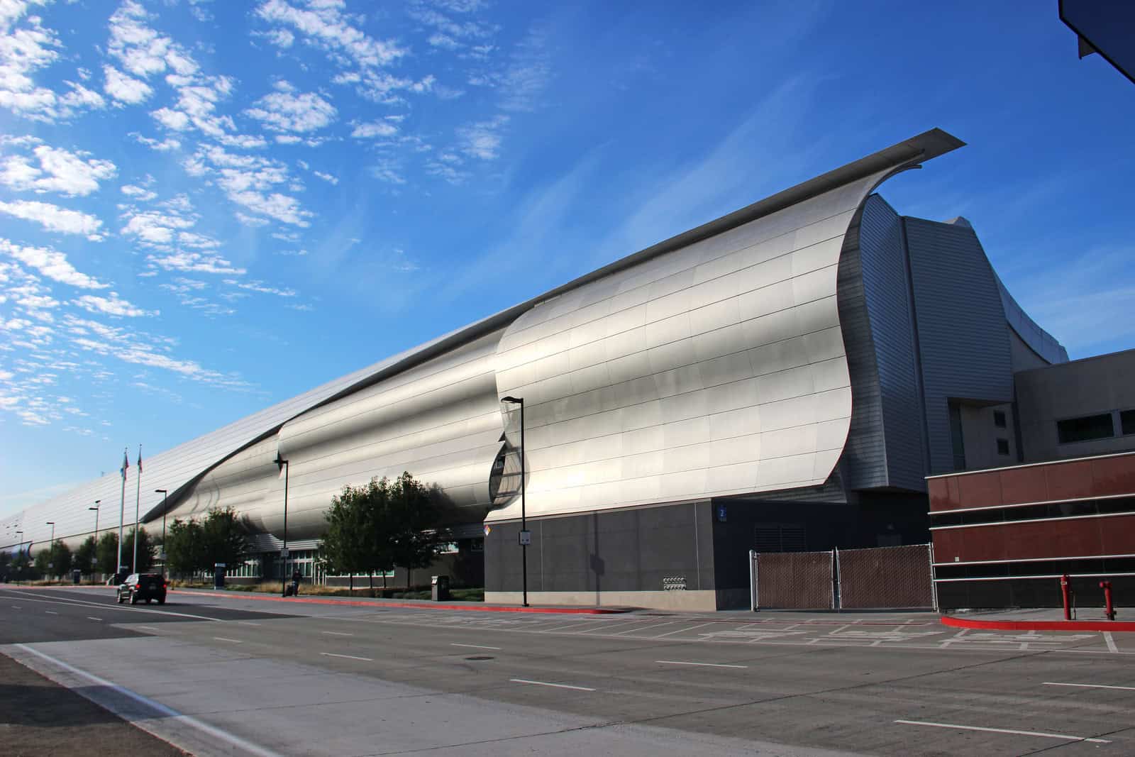 Large curved metallic building stretches along a quiet city street, its smooth silver panels catching sunlight under a bright blue sky with scattered clouds and a few trees.