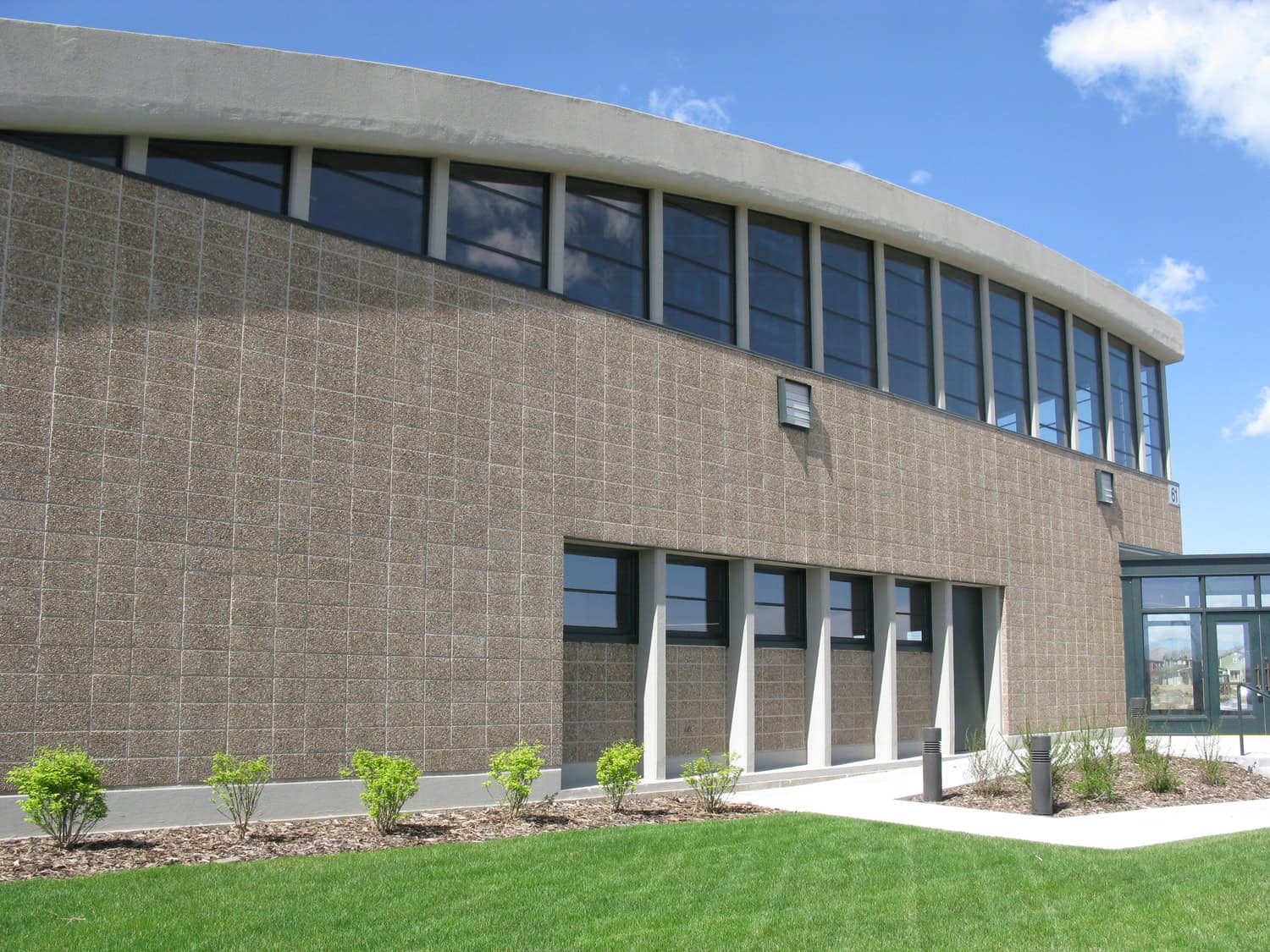 Institutional building facade featuring a curved white roof and upper and lower rows of windows; beige textured block wall faces a manicured lawn, shrubs, concrete walkway. Text: "61".