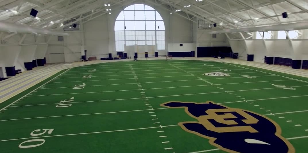 Indoor artificial-turf football field sits empty, marked with yard lines and a large CU logo, inside a white arched practice dome with a surrounding running track and big arched windows.

Text: "10", "20", "30", "40", "50", "PAC-12", "CU"