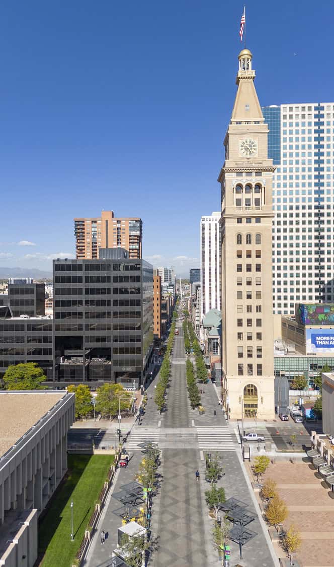 Clock tower stands beside a pedestrian-lined avenue, flanked by office buildings and trees, overlooking a long downtown street beneath a clear blue sky.