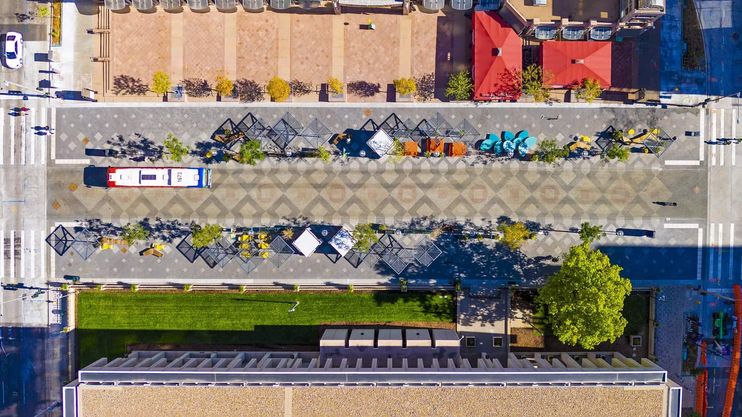 A city bus (number 1673) parked beside a central patterned pedestrian promenade lined with geometric seating, tables, trees and red-roofed kiosks between crosswalks and an adjacent green lawn.