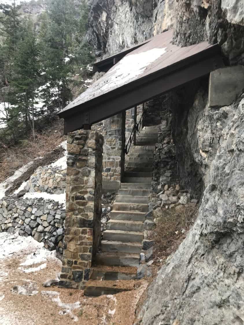 Stone stairway ascends alongside a rocky cliff, sheltered by a sloped metal roof supported by stone pillars; evergreen trees and patches of snow surround the narrow mountain path.