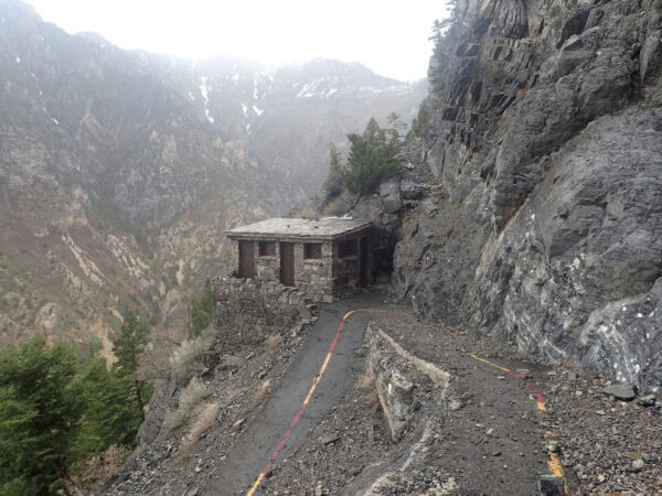 A small stone cabin clings to a narrow mountainside path, flanked by steep rocky cliffs on the right and a deep, misty canyon with sparse trees on the left.