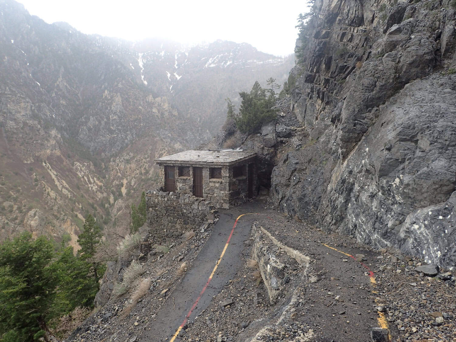 A small stone cabin clings to a narrow mountainside path, flanked by steep rocky cliffs on the right and a deep, misty canyon with sparse trees on the left.