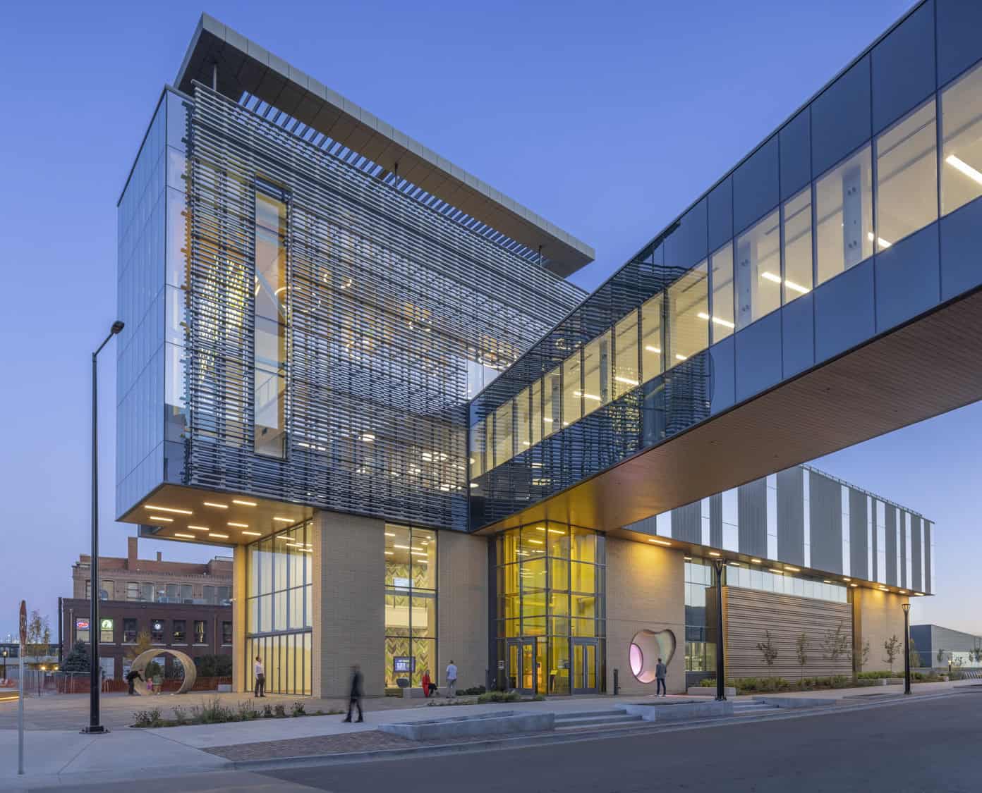 Contemporary glass-and-brick civic building displays a suspended glass skybridge and louvered facade, glowing interior lights illuminating an urban sidewalk at dusk with a few pedestrians passing by.