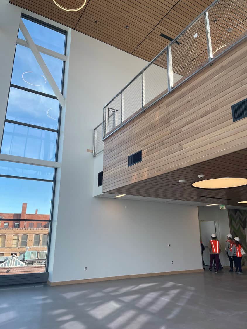 Wood-clad mezzanine overlooks empty atrium; tall floor-to-ceiling windows reveal brick buildings outside. Three people in hard hats and orange vests stand by a doorway labeled "EXIT".