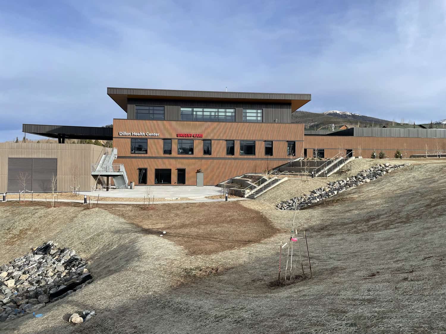 Dillon Health Center building sits on a gentle grassy slope with stairways leading to entrances; surrounded by rocky drainage, young trees, and distant snow‑capped hills under a clear blue sky. Text visible: "Dillon Health Center" and "URGENT CARE".