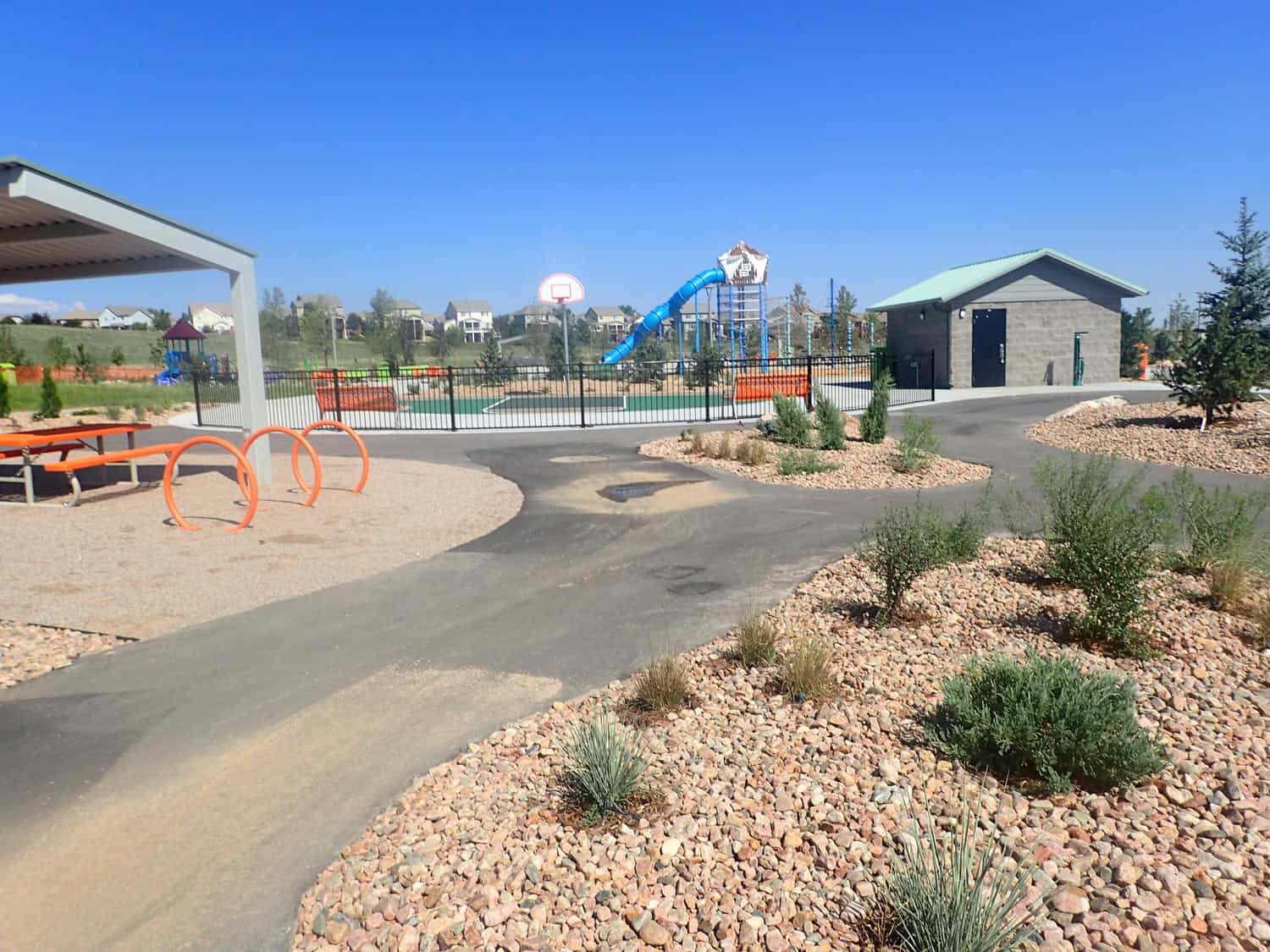 A tall play tower with spiraling blue tube slide descends into a fenced playground beside a basketball hoop and picnic shelter, with paved paths and rock-mulched landscaping under clear sky.