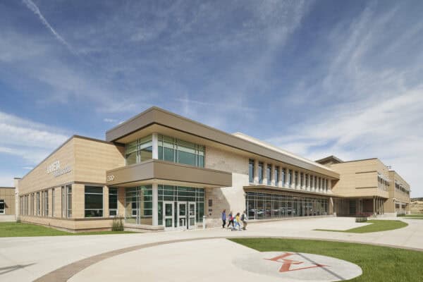A modern school building; a small group of students walk toward the glass entrance amid a paved courtyard and lawns under a broad blue sky.

Visible text: "LA VETA PUBLIC SCHOOLS", "290", stylized "LV" on pavement.