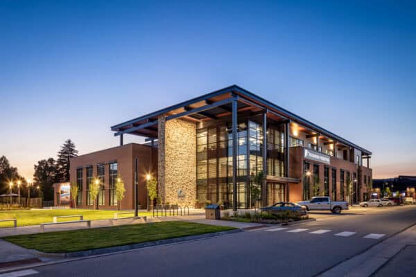 Modern brick-and-glass building (sign: "STEELHEAD BUILDING") lit at dusk, showcasing a glass atrium and stone column, facing a landscaped plaza with benches and parked cars on a quiet street.