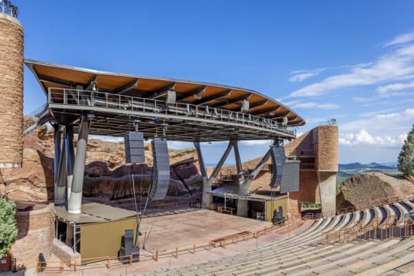Outdoor amphitheater stage supported by a steel roof and hanging speakers, standing ready for a performance, set into red rock formations with terraced concrete seating and a clear blue sky.