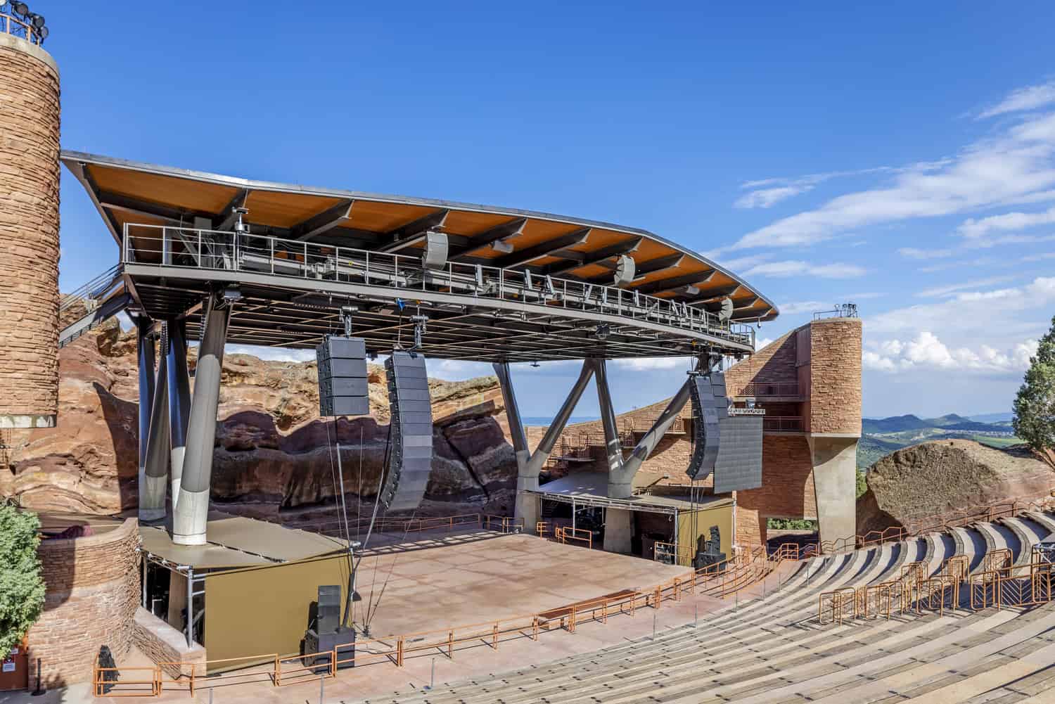 Outdoor amphitheater stage supported by a steel roof and hanging speakers, standing ready for a performance, set into red rock formations with terraced concrete seating and a clear blue sky.