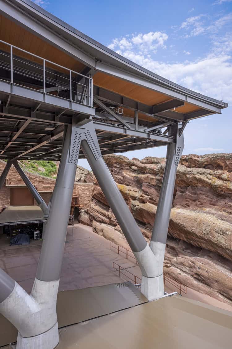 V-shaped steel support columns supporting a wide cantilevered roof above an empty concrete stage, framed by layered red sandstone cliffs and a partly cloudy blue sky.