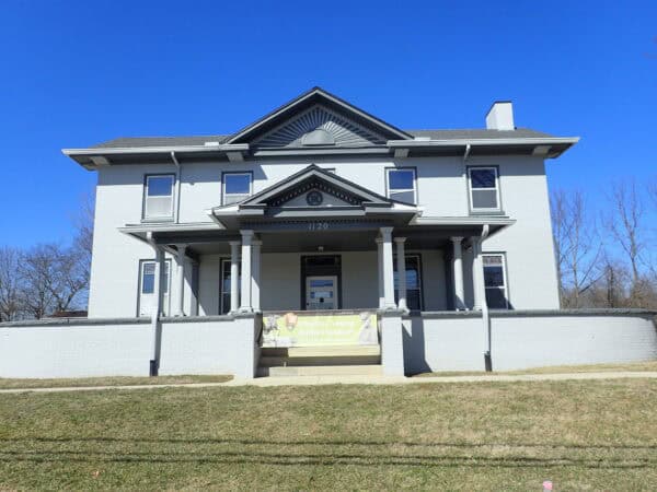 Two-story gray brick house (1120) stands centered on a grassy lawn under a clear blue sky, with a columned front porch and banner reading: "Charles Young Buffalo Soldiers National Monument."