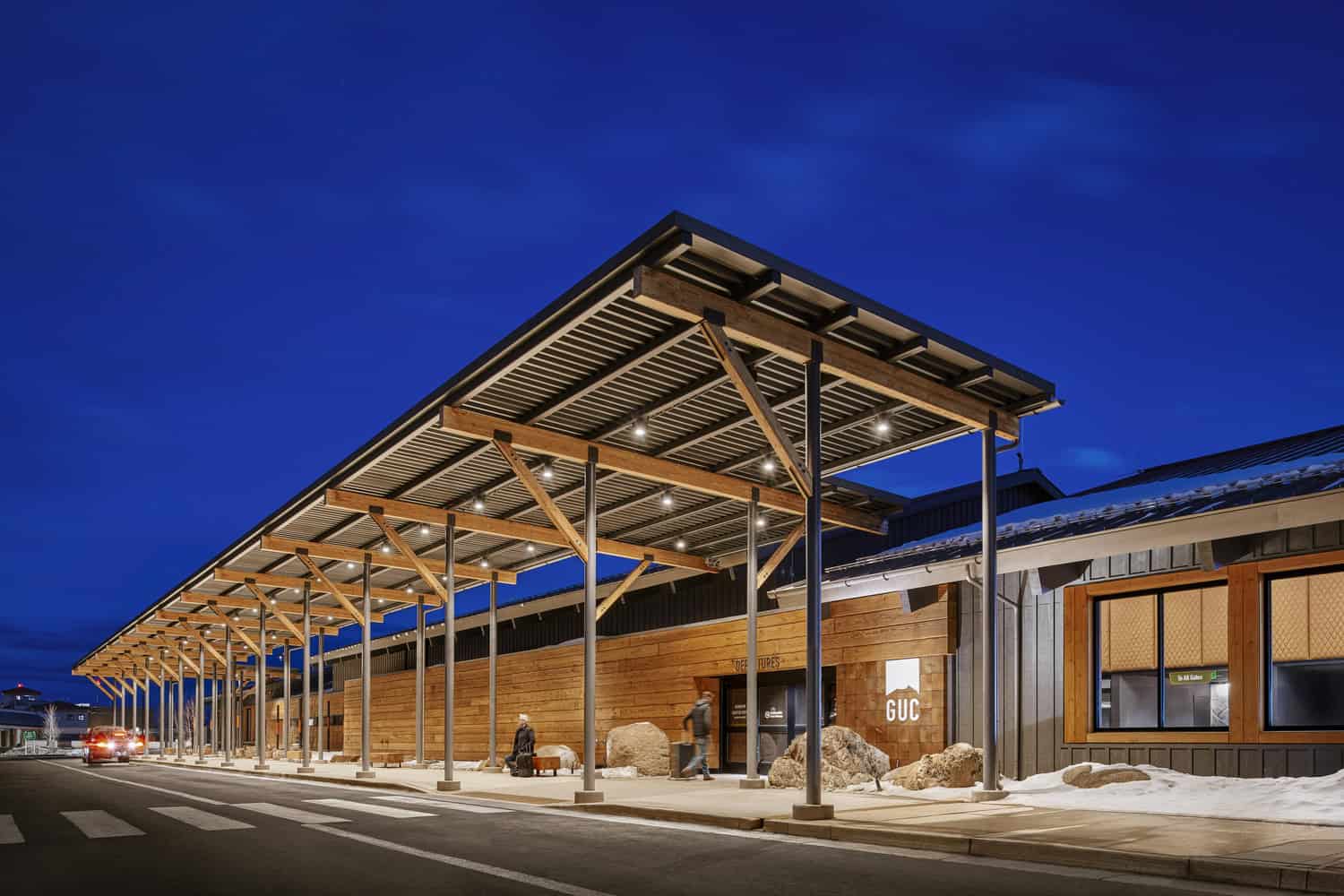Wooden airport terminal canopy illuminated at night, with passengers walking and sitting beneath it by a snow-lined curb, crosswalk and a stopped car under a deep blue sky.

Text visible: "GUC" ; "DEPARTURES" ; "To All Gates"