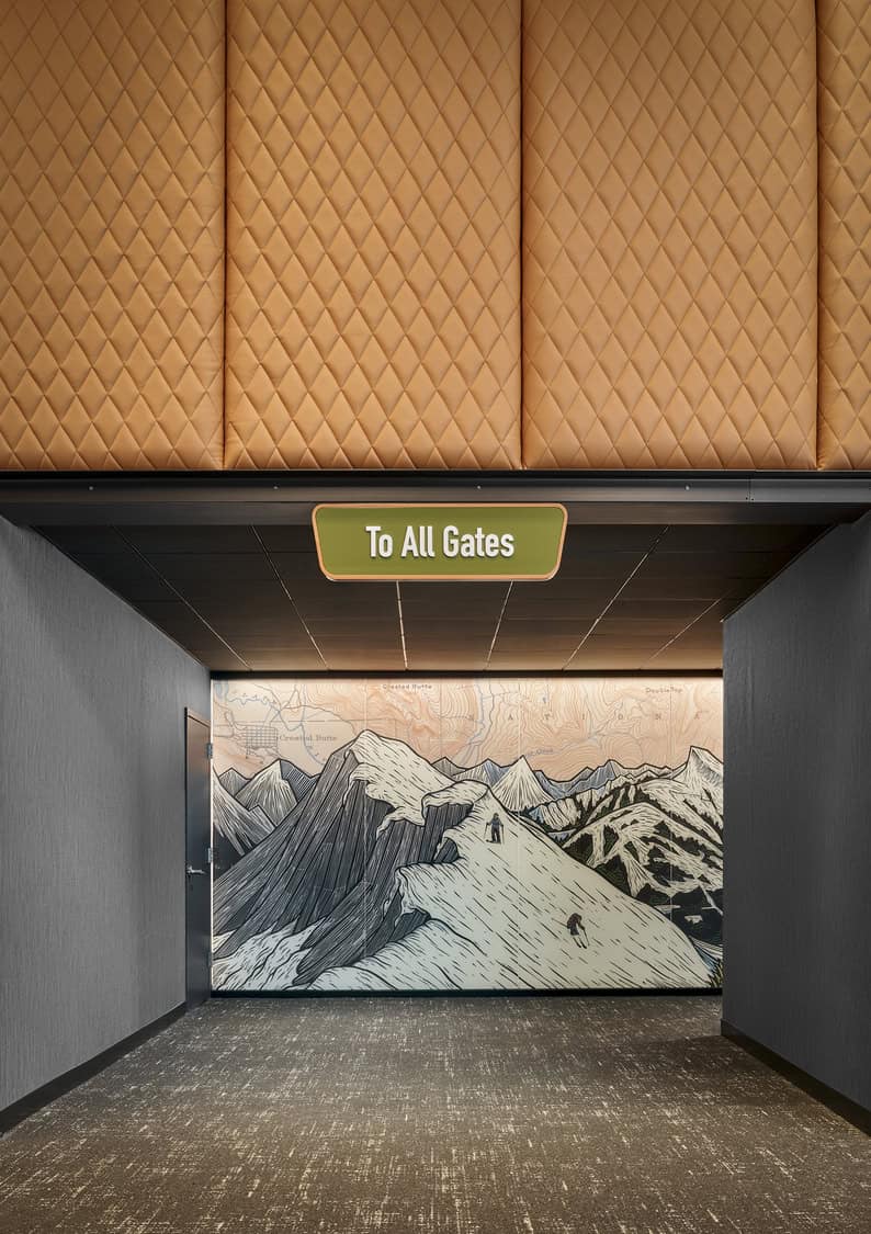 Green overhead sign reading "To All Gates" hangs above a carpeted corridor, framed by quilted tan panels and leading to a large stylized mountain mural on the far wall.