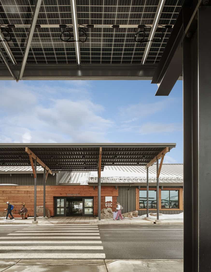Covered terminal entrance framed by a solar-panel canopy, people walking and waiting at a zebra crossing, wooden-clad facade and snowy ground under a blue sky. Text: "GUC"; "DEPARTURES".