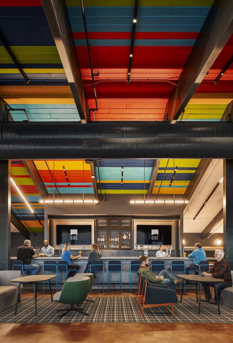 Bar beneath a vibrant multicolored striped ceiling; patrons sit at the counter and lounge while bartenders serve drinks in an industrial space with exposed ducts — two TV screens read "GUC".