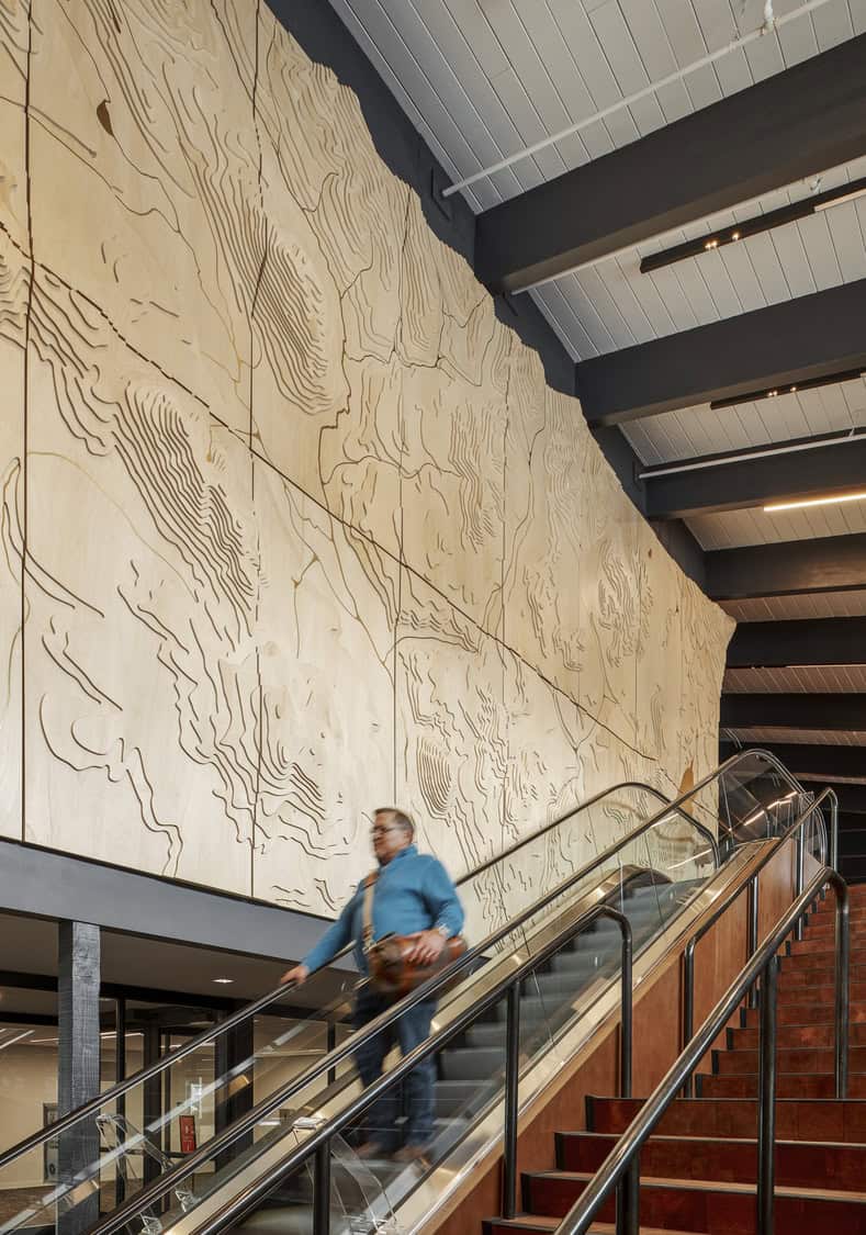 Large carved plywood relief wall depicting topographic contour lines stretches above an indoor escalator; a blurred person rides down, steel handrails and exposed ceiling beams frame the modern lobby.