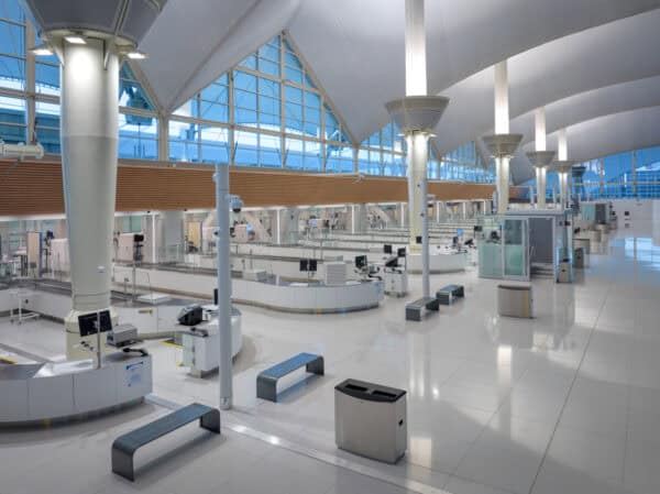 Airport check-in counters sit empty, arranged in curved islands with monitors and benches, beneath vaulted white ceilings and large blue‑tinted windows over a glossy tiled floor.