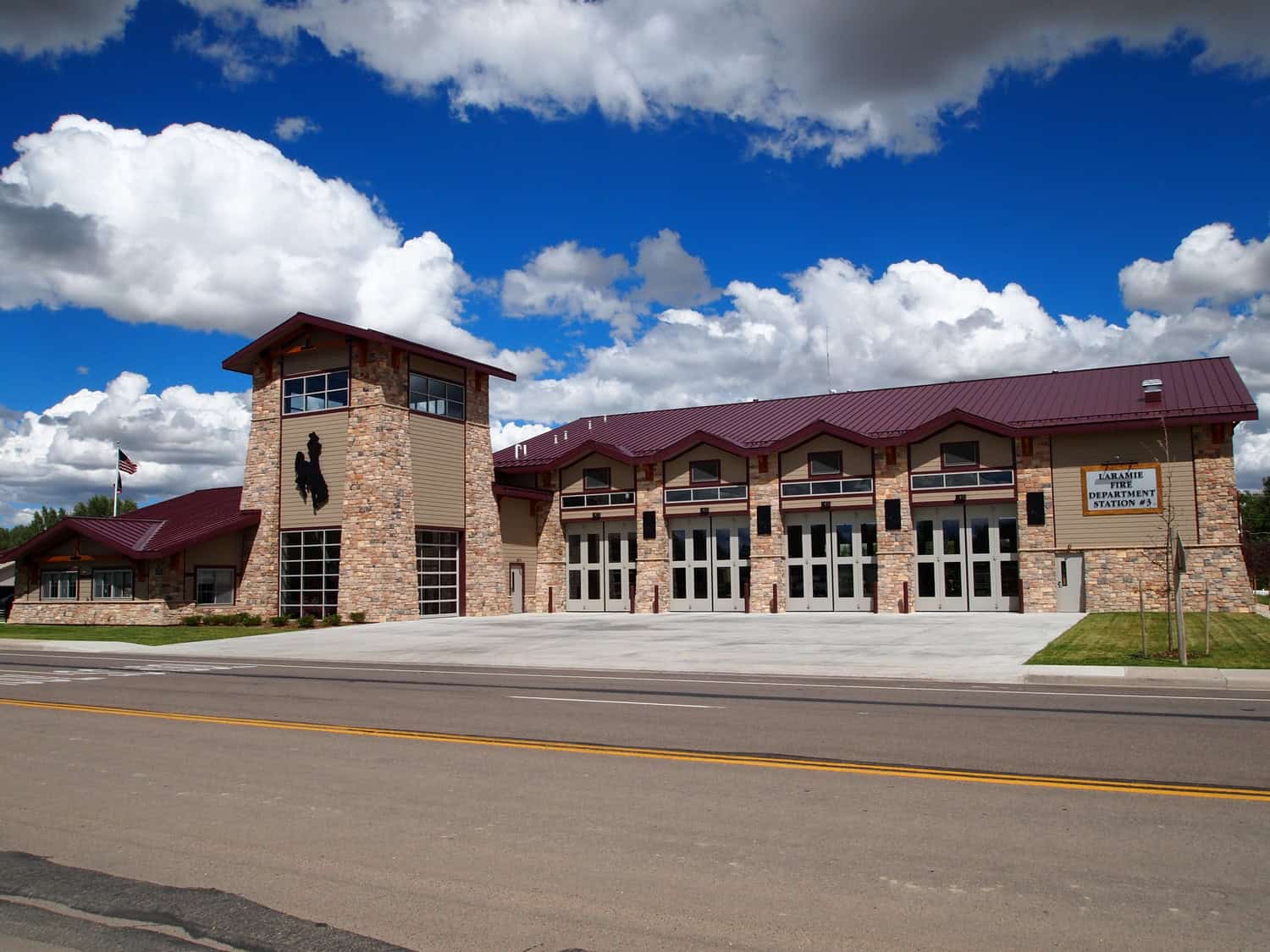 Fire station with stone tower and silhouette emblem, several closed bay doors, American flag nearby, concrete apron and empty street, under vivid blue sky with large white clouds.

Text in image: "LARAMIE FIRE DEPARTMENT STATION #3"