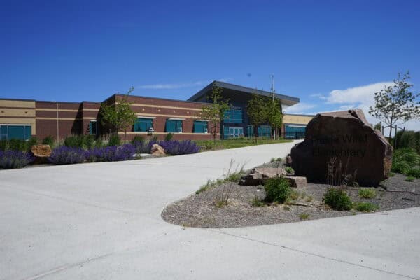 A single-story brick elementary school sits behind a large engraved boulder, with purple flower beds and young trees lining a wide concrete driveway under a clear blue sky.

Text: "Prairie Wind Elementary" (appears on the entrance boulder and building).