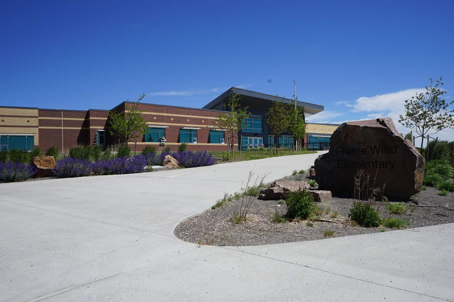 A single-story brick elementary school sits behind a large engraved boulder, with purple flower beds and young trees lining a wide concrete driveway under a clear blue sky.

Text: "Prairie Wind Elementary" (appears on the entrance boulder and building).