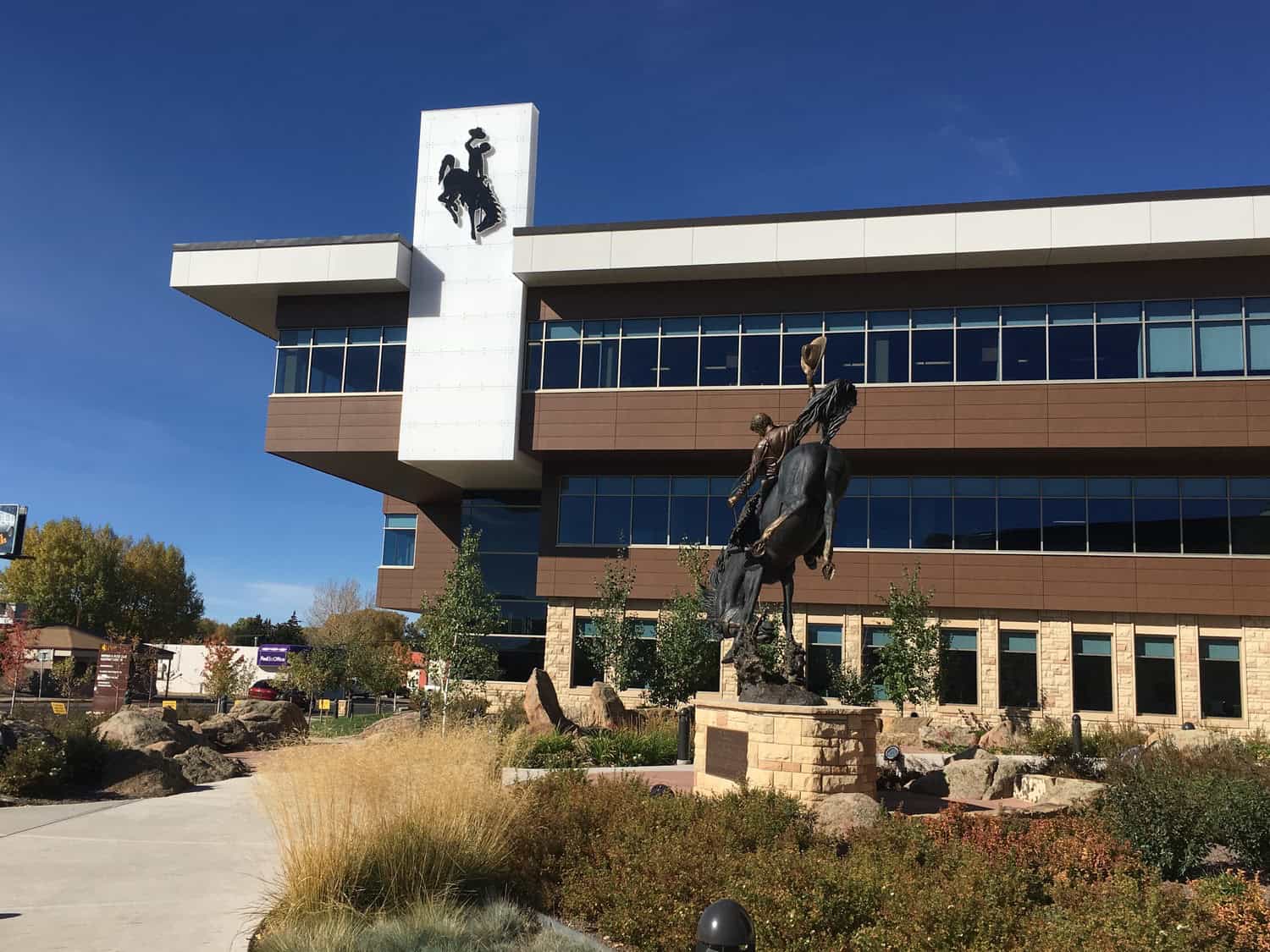 Bronze statue of a cowboy raising his hat while riding a rearing horse, set on a stone pedestal in a landscaped plaza before a brown-and-white building under a clear blue sky.