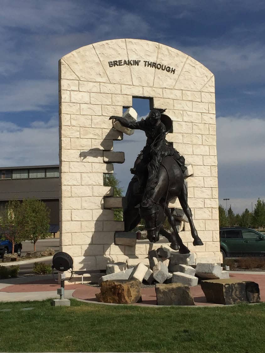Bronze cowboy on a rearing horse bursts through a stone wall reading BREAKIN' THROUGH, surrounded by rubble in an outdoor plaza with grass, cars and a building under blue sky.