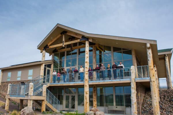 Lodge-style building with large glass façade and log posts, hosting a row of people standing along the second-floor balcony, overlooking rocky landscaping beneath a pale blue sky.