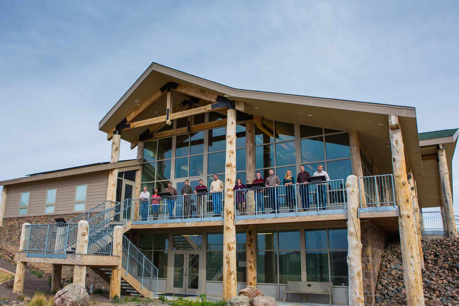 Lodge-style building with large glass façade and log posts, hosting a row of people standing along the second-floor balcony, overlooking rocky landscaping beneath a pale blue sky.