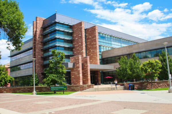 A brick-and-glass academic building stands above steps as a person climbs them on a sunny campus plaza with trees and outdoor tables. Sign: "Behavioral Sciences 410 Pitkin Street".