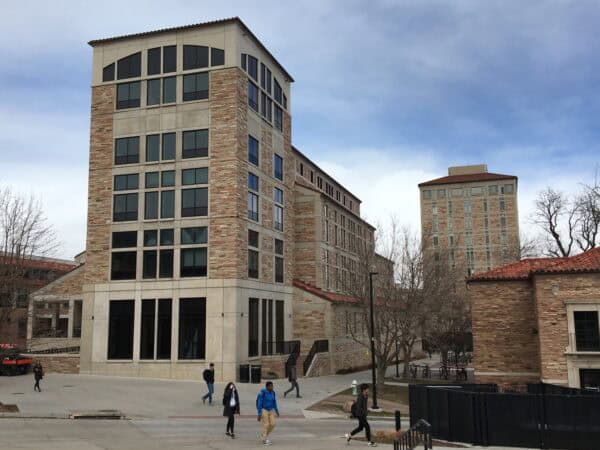 Multi-story stone-and-glass academic building towers over a concrete plaza; students walk across the plaza, with leafless trees and additional campus buildings surrounding under a cloudy sky.