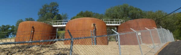 Three large rust-colored cylindrical storage tanks stand on a gravel slope, connected by a white catwalk; a barbed-wire chain-link fence separates them from a road beneath a clear blue sky.